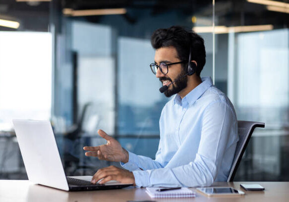 Indian young man wearing a headset conducts a webinar, online conference sitting at a laptop in the office.
