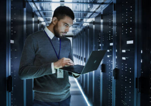 IT Technician Works on a Laptop in Big Data Center full of Rack Servers. He Runs Diagnostics and Maintenance, Sets up System.